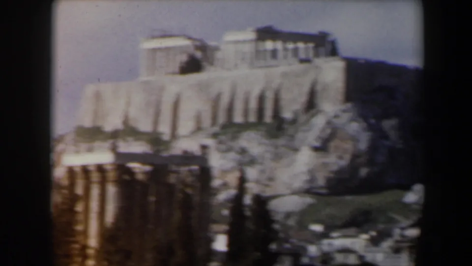 1960s 8mm travel frame showing the Acropolis area from a distance, with pale marble structures above rocky slopes and foreground ruins.