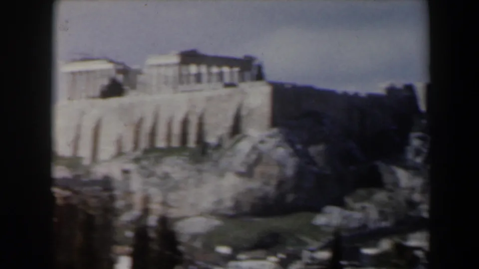 1960s home-movie view of a pale classical landmark on a rocky ledge, with dark trees, ruins, and surrounding hillside visible.