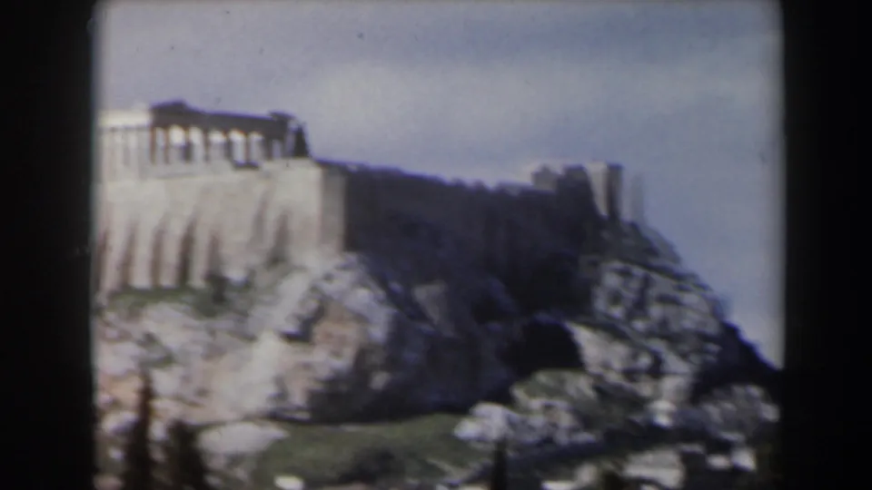 Faded 8mm frame showing part of the Acropolis area on a rocky ridge, with pale stone architecture, shadowed hillside, and open sky.