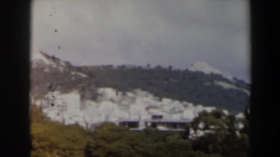 Wide 8mm travel view of a hillside city area below a rocky summit, with buildings and trees visible.