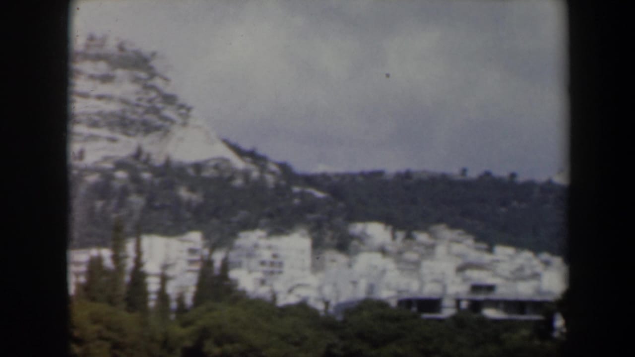 Snowy mountain slope with pines, distant white buildings, and an overcast sky.