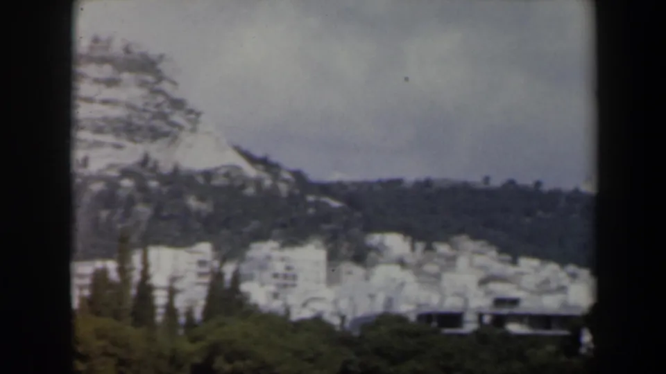 1960s Israel 8mm frame showing a snow-dusted mountain slope with pines and distant white buildings below the ridge.