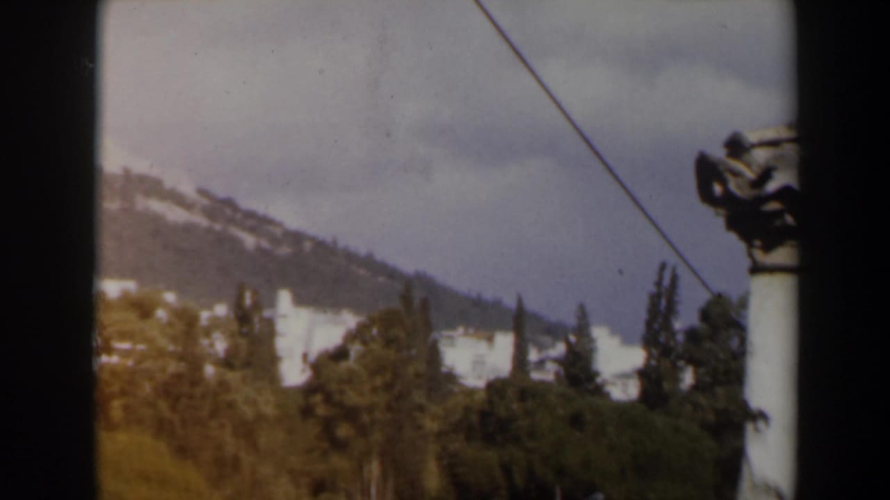 Mountain slope with white buildings, trees, clouds, and a diagonal wire.