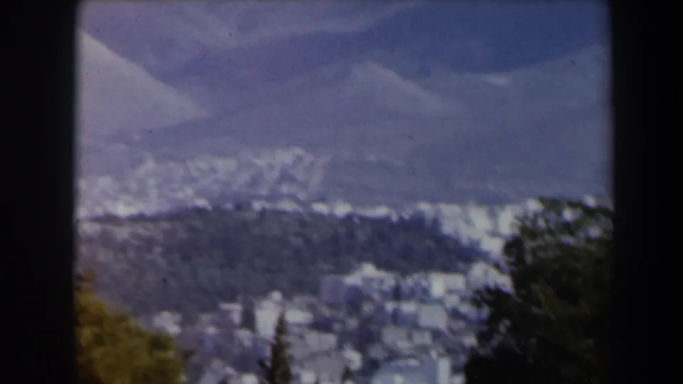 1960 Israel 8mm frame showing distant white hillside buildings, dark trees, and a hazy mountain backdrop.