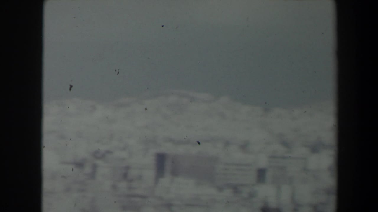 1960 Israel home-movie frame with white hillside structures and a muted mountain backdrop.