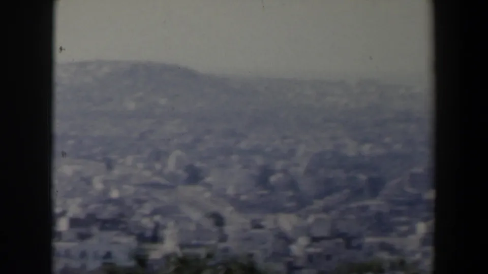 Archival 8mm frame from Israel in 1960 showing a hazy city spread across hills, partly framed by bushes.