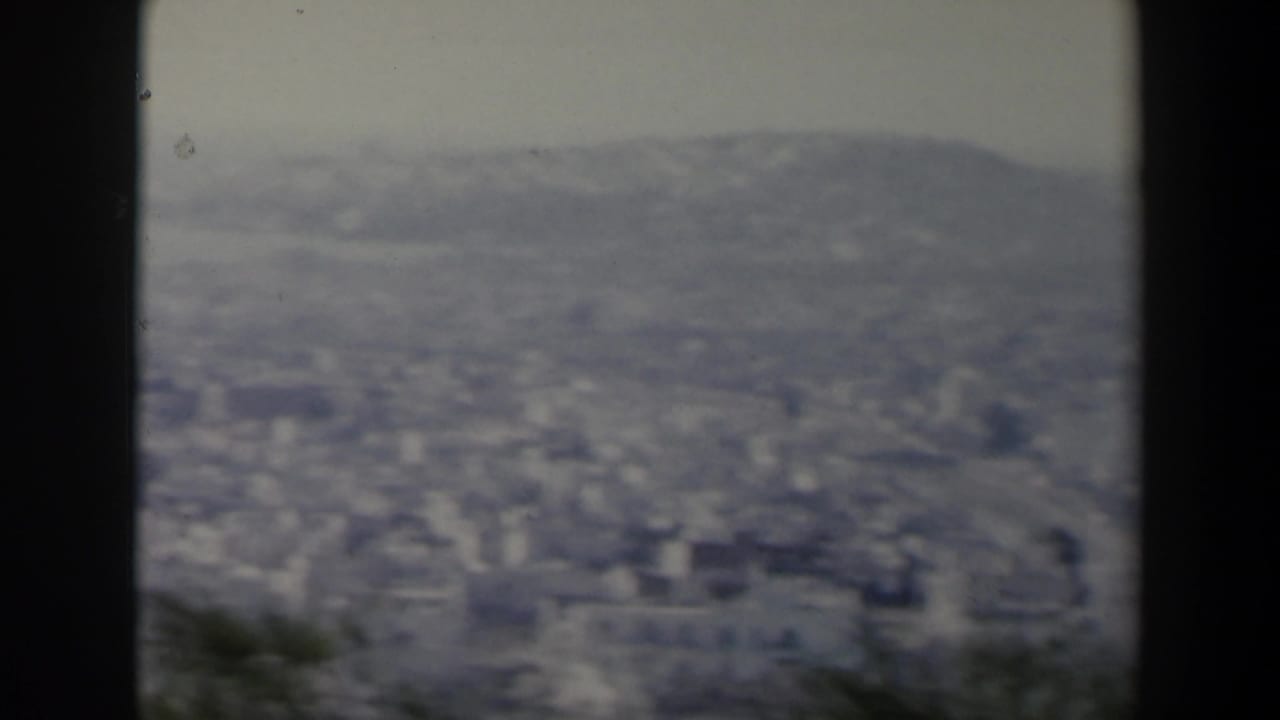1960 8mm view in Israel showing a distant city below rolling hills, framed by dark gate edges and soft focus.