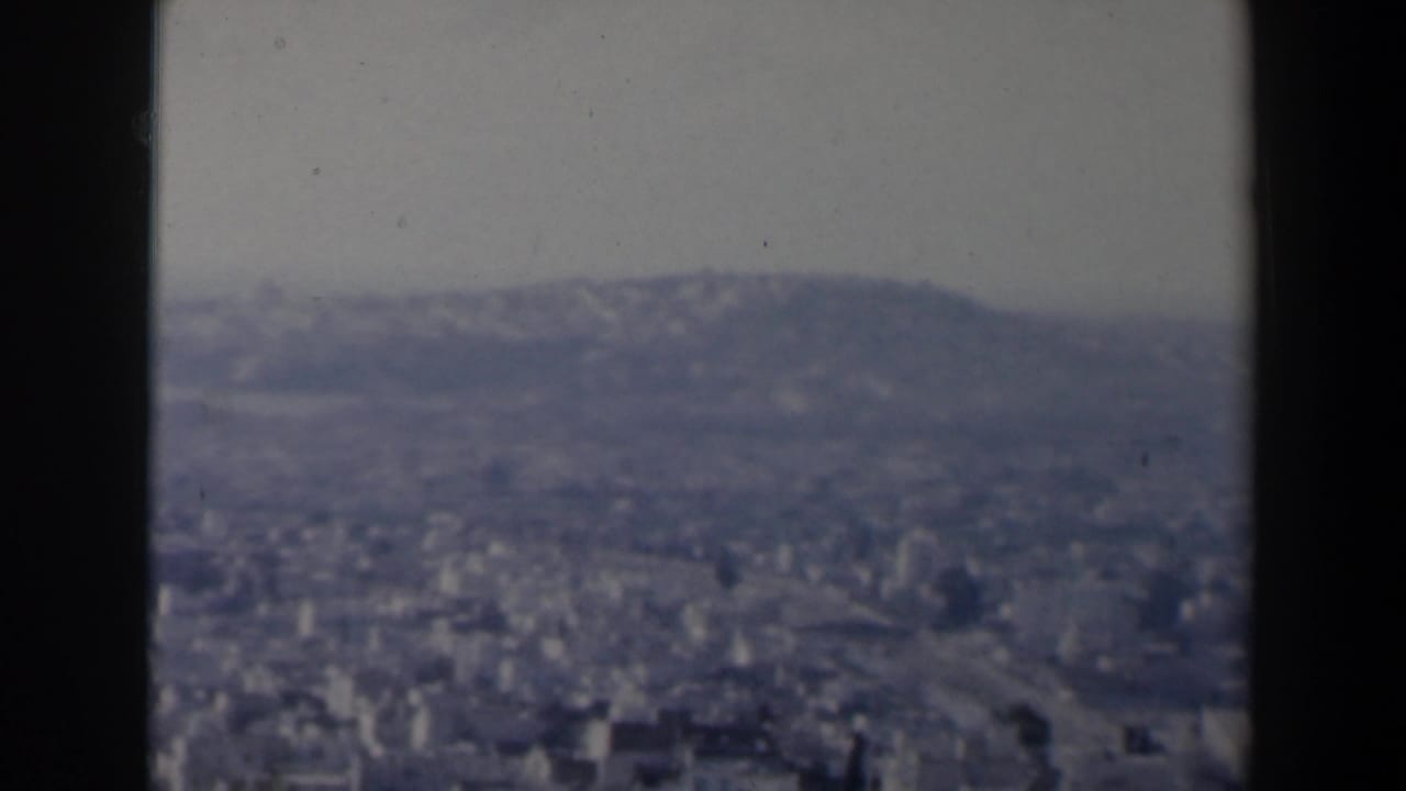 1960 Israel home-movie frame showing a hazy city panorama from a high viewpoint, partly framed by dark vertical edges.