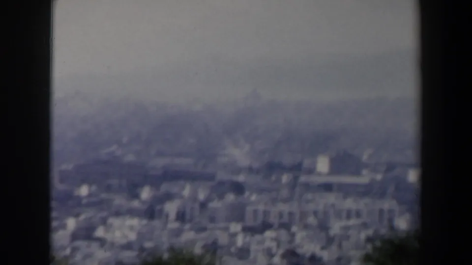 Hazy 1960 8mm view over an Israeli city, showing dense pale buildings, distant terrain, and dark foliage at the frame edge.