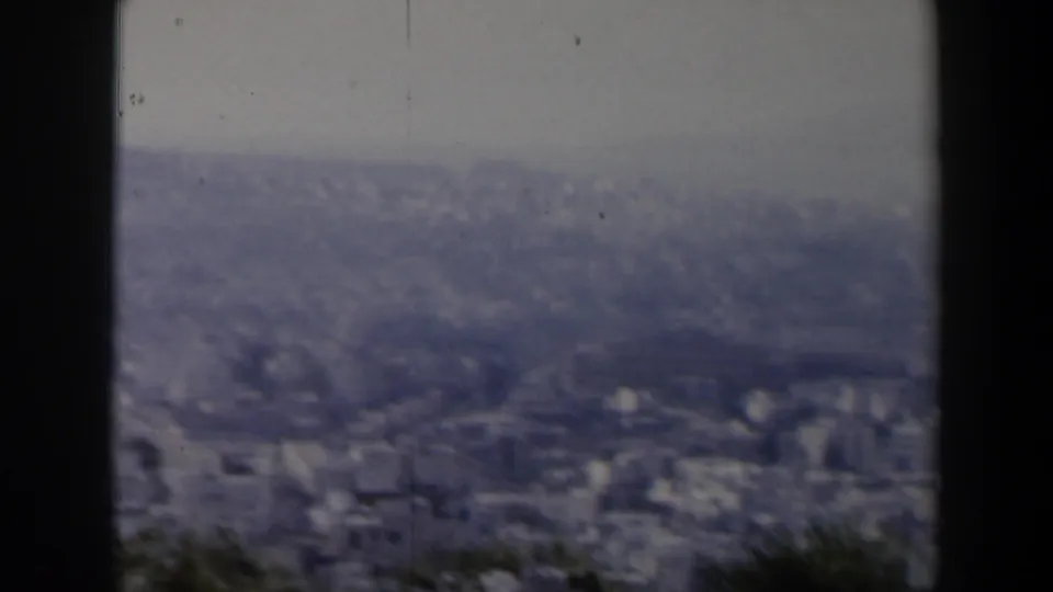 1960 Israeli home-movie frame showing a broad hazy city view, foreground vegetation, and dark film or tree edges around the scene.