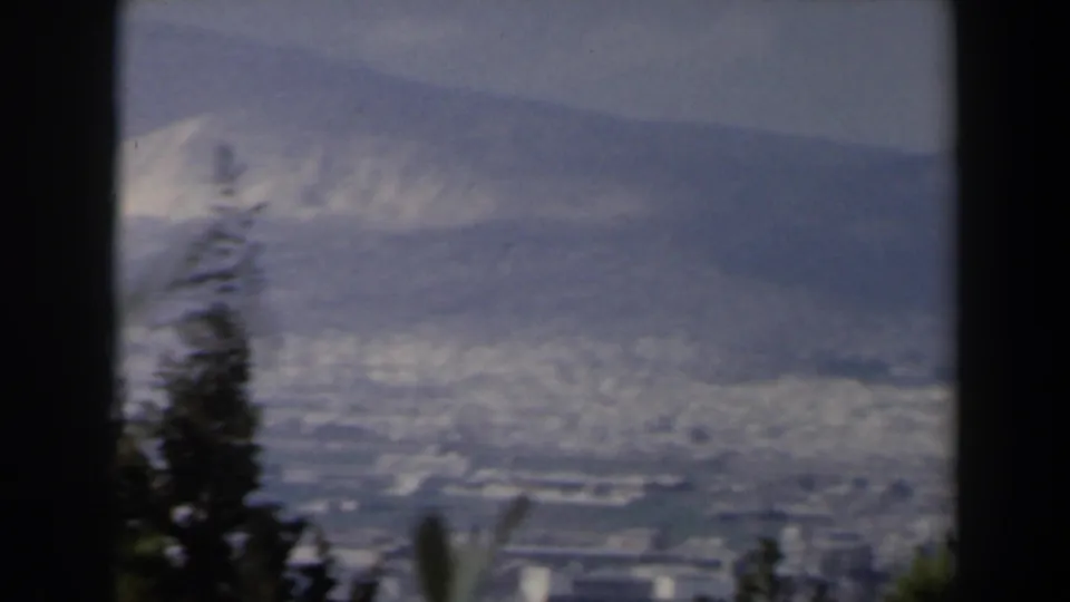 1960 Israel hilltop panorama showing trees in the foreground, a hazy urban area below, and mountains along the horizon.