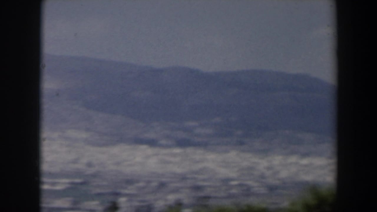 Distant valley city below hazy mountains, framed by dark 8mm borders and foreground greenery.