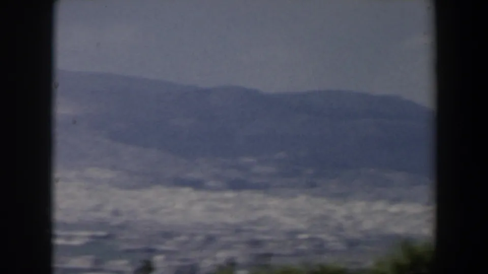 1960 Israel 8mm view of a distant city spread across a valley beneath hazy mountains, seen past dark frame edges and shrubs.