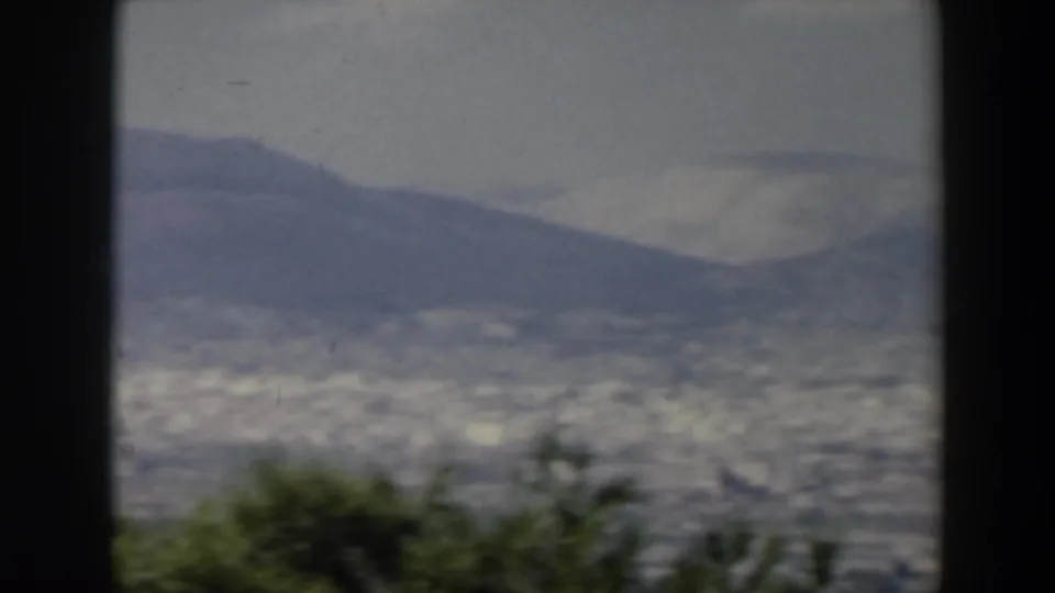 1960 Israel home-movie frame showing a hazy valley city beneath mountains, with dark 8mm borders and low foreground trees.