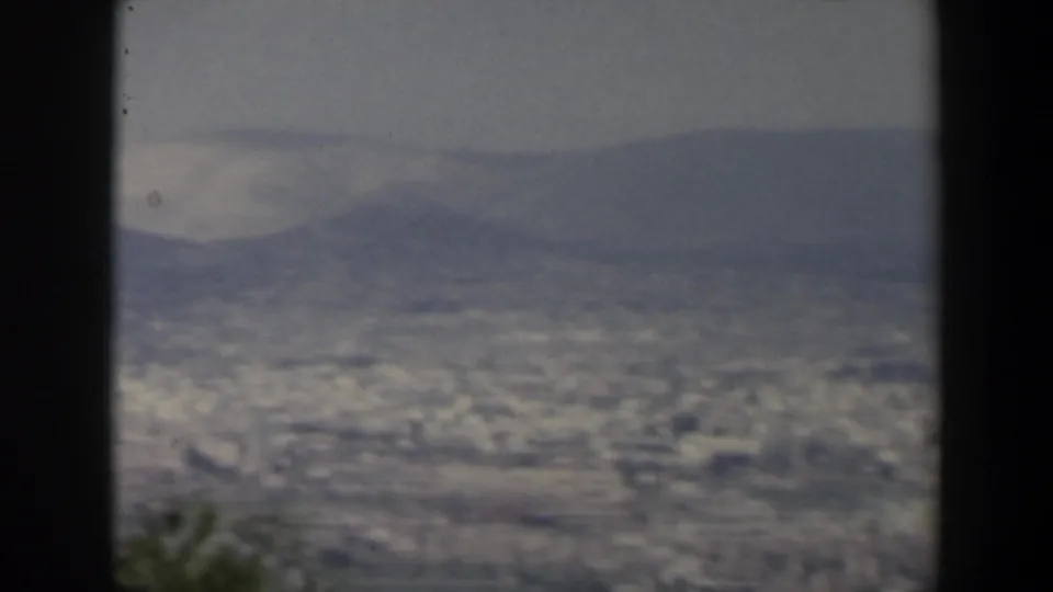 A wide 1960 Israel 8mm valley panorama with pale city forms, hazy mountains, and blurred greenery at the lower edge.