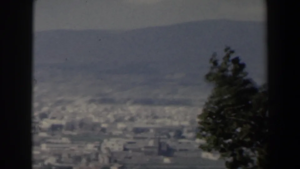 1960 Israel 8mm frame of a distant valley city below mountains, with a tree on the right and dark film borders enclosing the view.