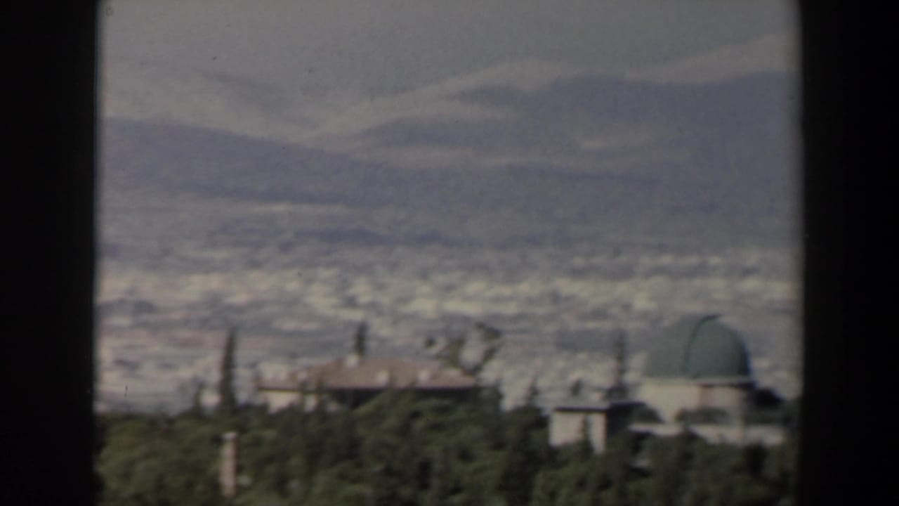 Distant city and mountains seen past dark vertical framing, with a green-domed building on a wooded ridge.