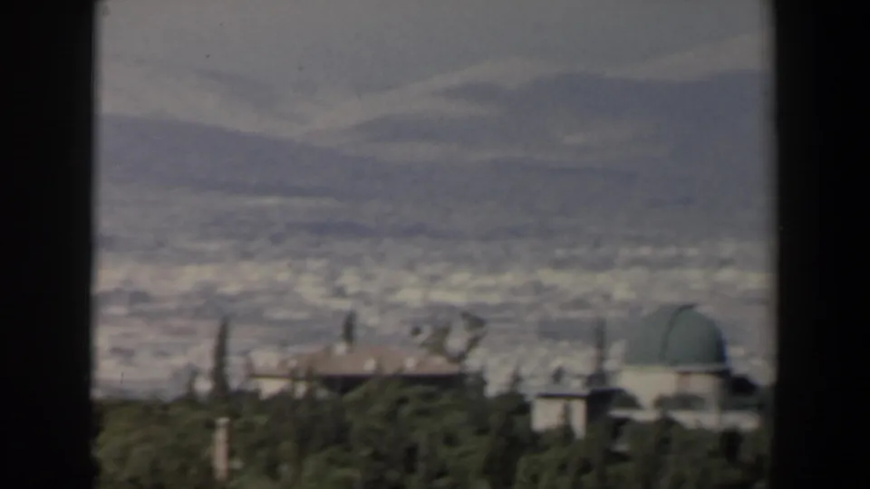 1960 Israel 8mm view of a hazy city below mountains, framed by dark foreground edges and a green-domed building near trees.