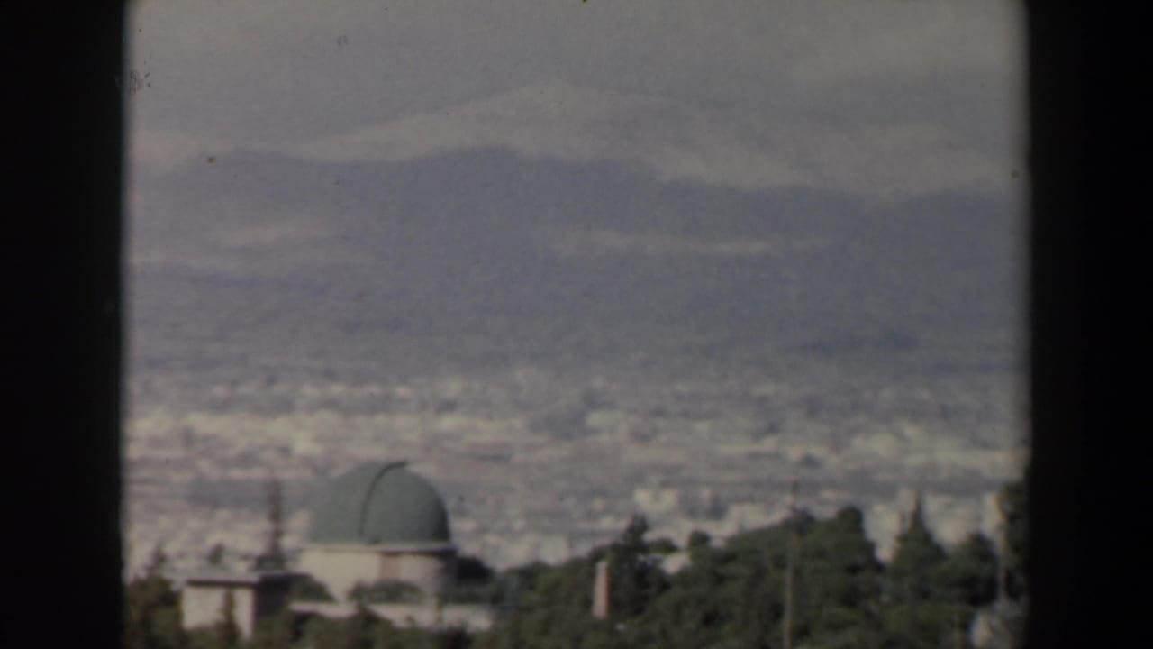 Elevated 1960 Israel home-movie frame showing a green-domed building, trees, distant city blocks, and hazy mountains.