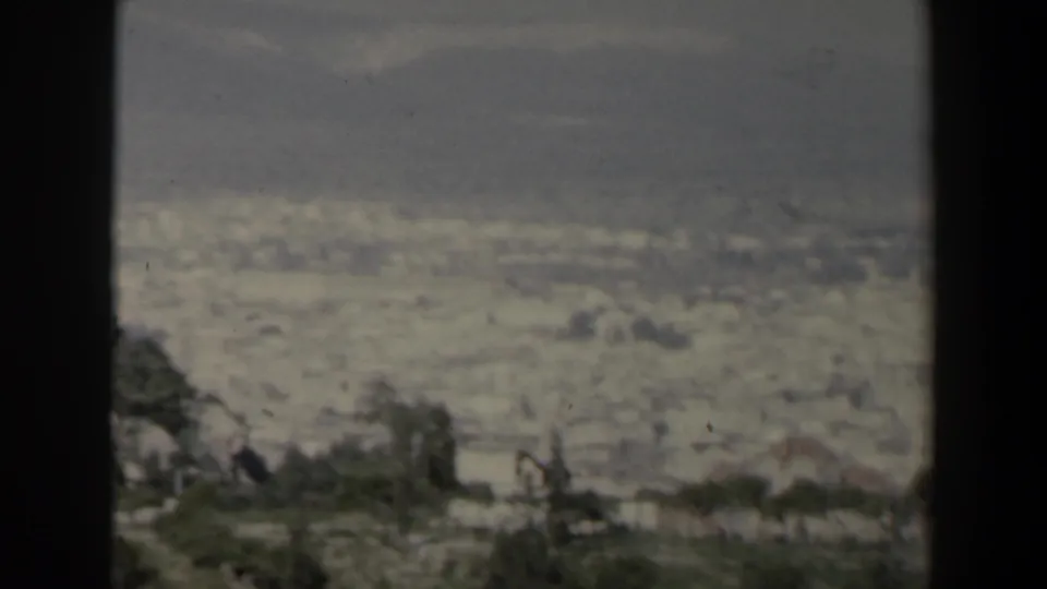 1960 Israel 8mm panorama of a sprawling city beneath mountains, with trees and dark foreground borders at the edge of the view.