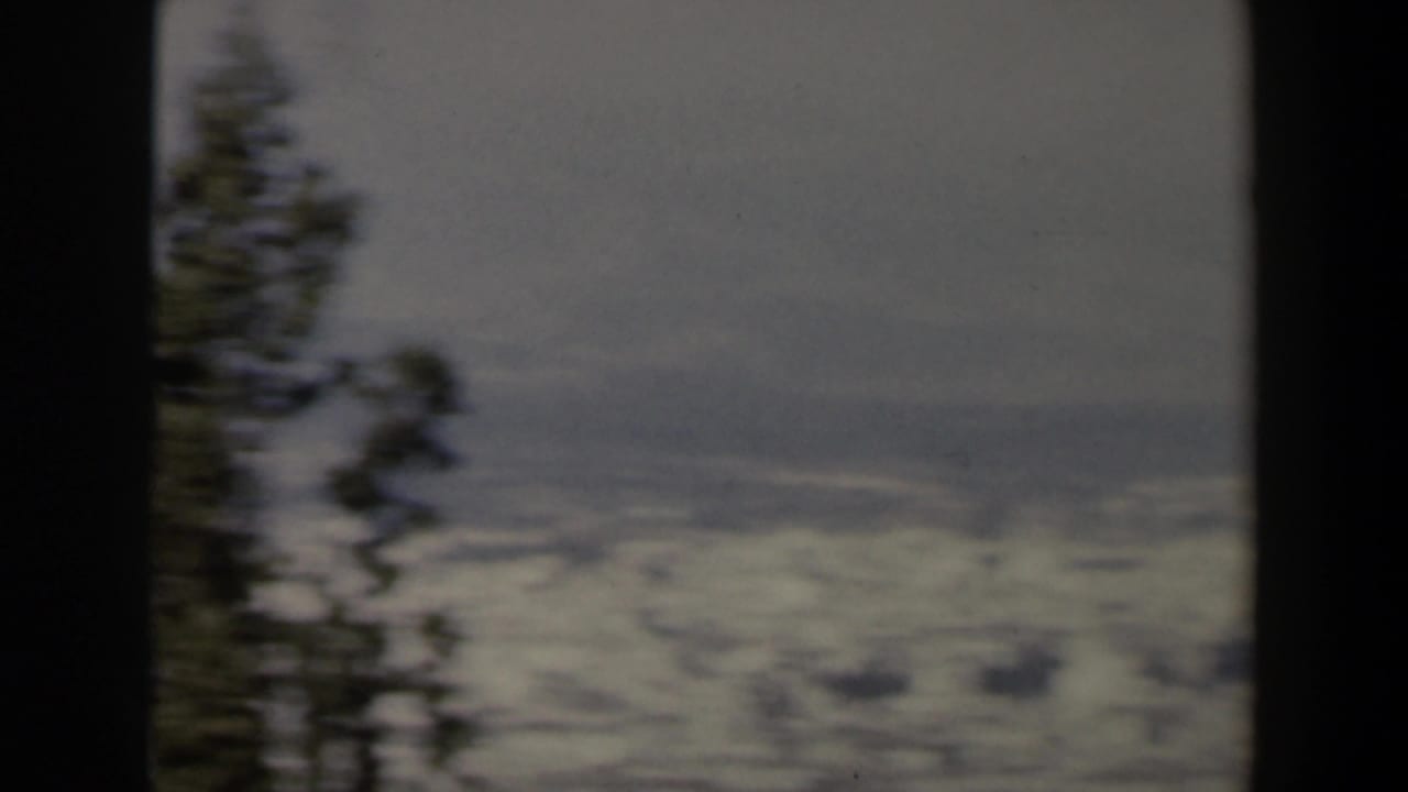 1960s Israel home-movie frame showing a snow-covered valley and hazy mountain slopes behind a dark foreground frame and conifer.