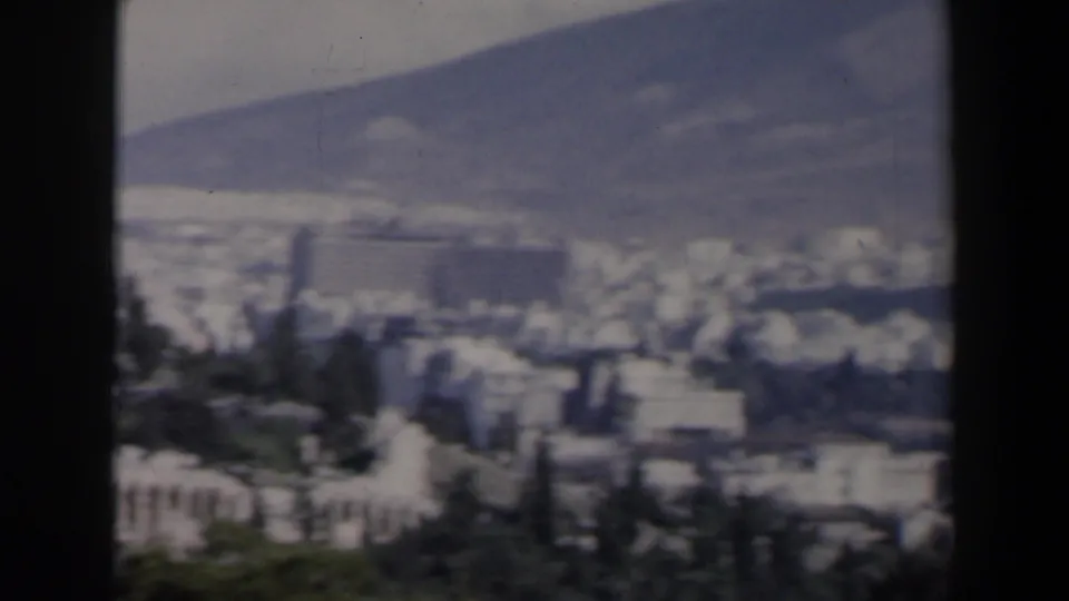 1960s 8mm view over Athens-like city buildings toward a hazy mountain backdrop, with trees in the foreground.