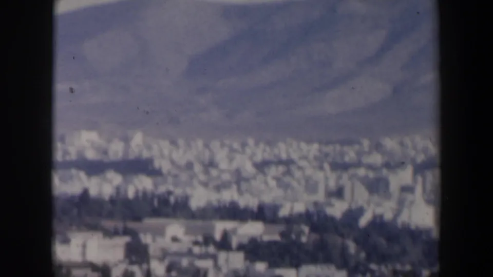 Wide 8mm city panorama, likely Athens based on clip context, with dense pale buildings and blue-gray mountains beyond.