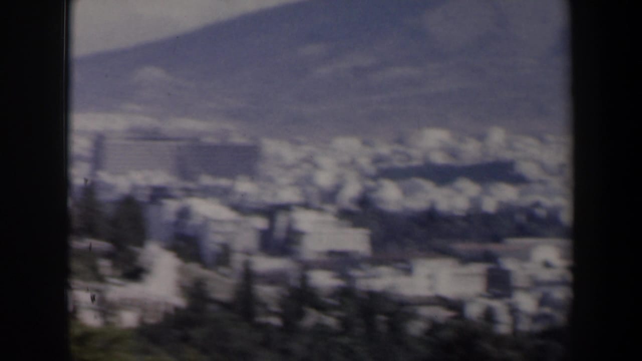 Distant city buildings below a hazy mountain range, seen through dark film-frame borders.