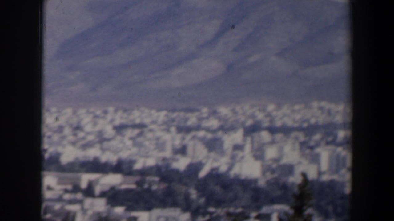 Pale city blocks and roads at the foot of mountains under a hazy sky.