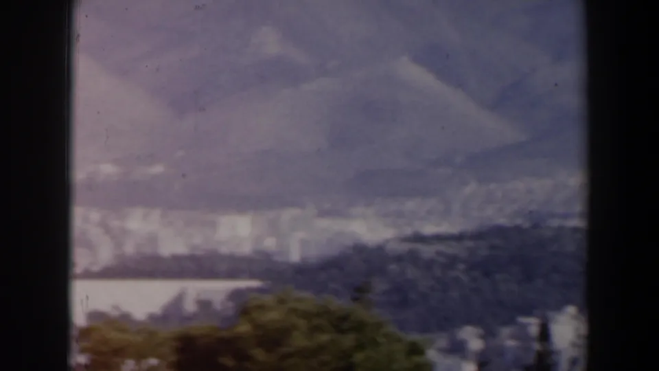 Faded 1960 8mm Israel city view with dark hillside, trees, pale buildings, and mountains beyond.