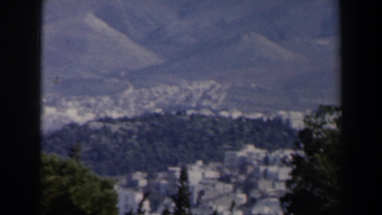 Forested overlook with a distant valley settlement and mountains beyond.