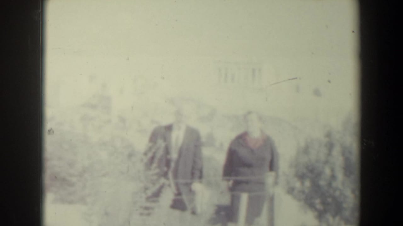 Two adults stand outdoors near a railing with trees in bright light.