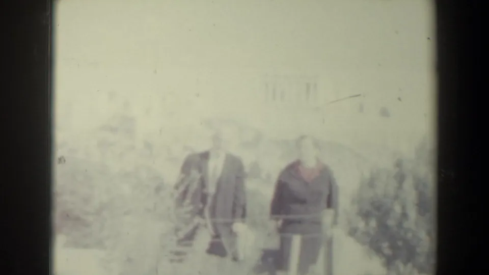In 1960s Israel home-movie footage, a man in a dark suit and a woman with a red collar stand beside trees near a railing.