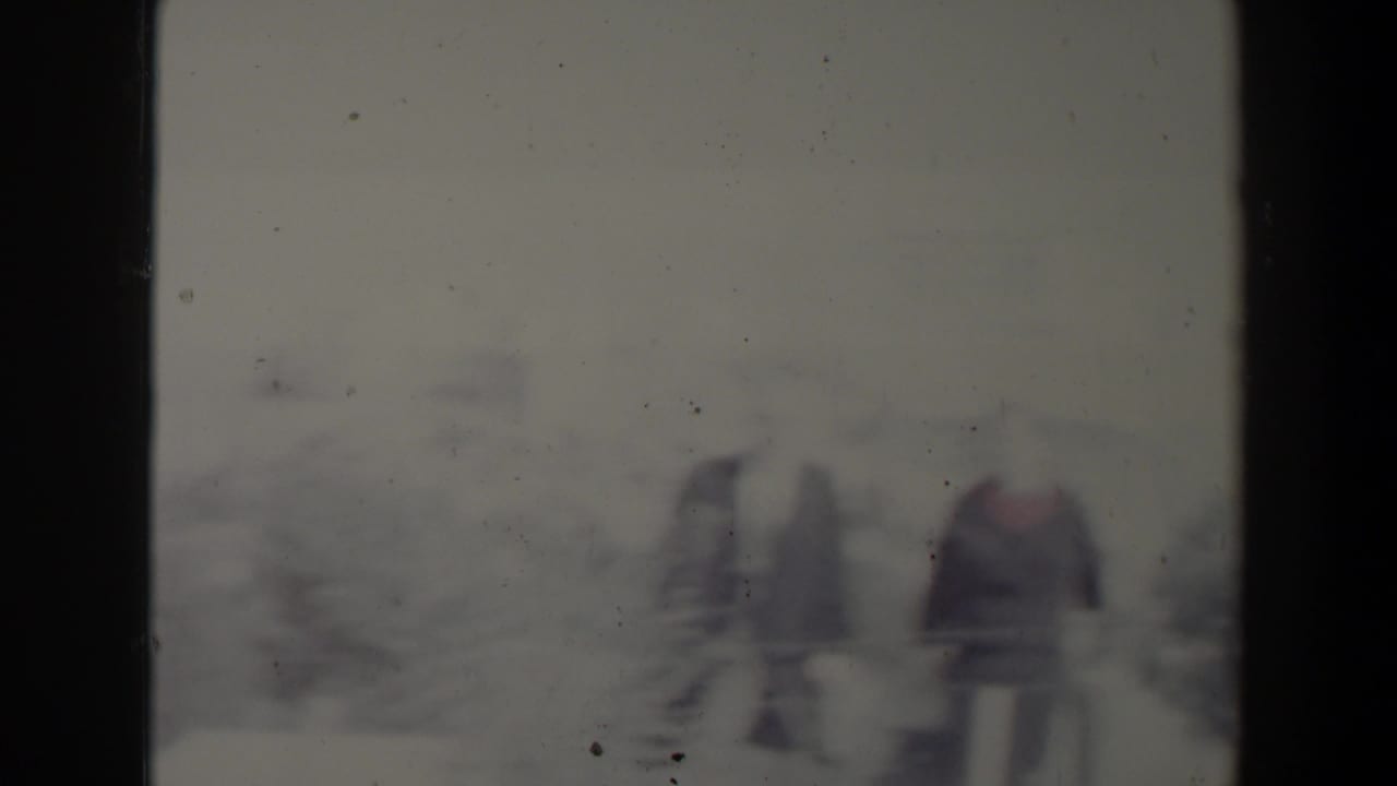Two blurred adults stand outdoors near bushes, seen through a dark 8mm frame border.