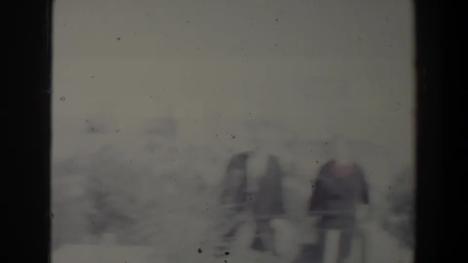 Two adults appear in a soft, grainy 1960 Israel home-movie frame, standing outdoors with bushes and pale ground behind them.