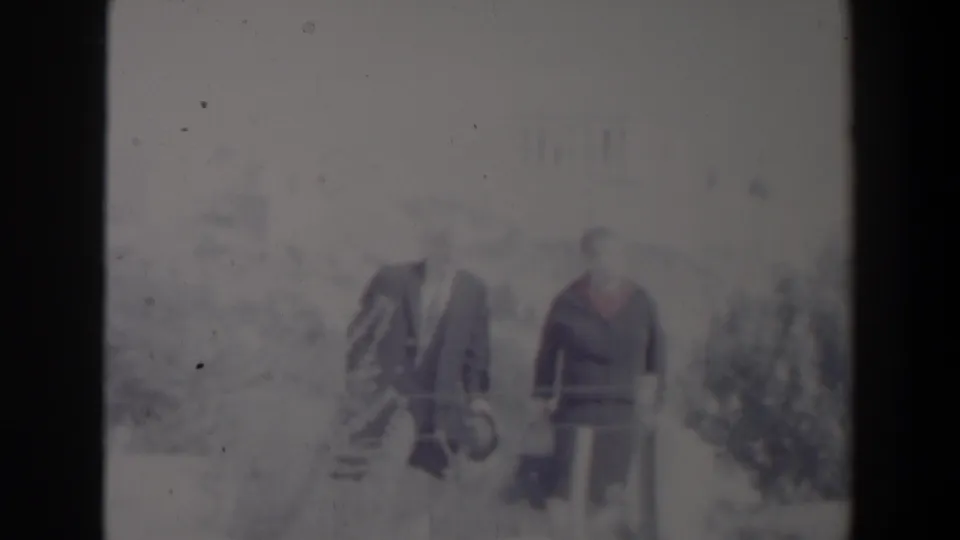 Two adults remain posed outdoors in the 1960 Israel home-movie footage, framed by low bushes and a pale, overexposed background.