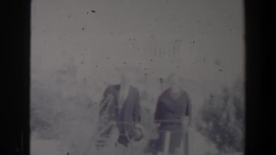 Two adults in period clothing walk outdoors in the Israel home-movie footage, with heavy fading and visible film specks.