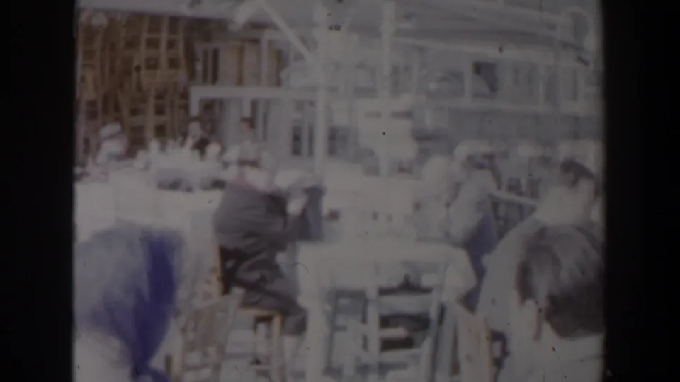 Community gathering scene in 1960 Israel, with adults seated at tables under a bright structure in soft, overexposed 8mm color.