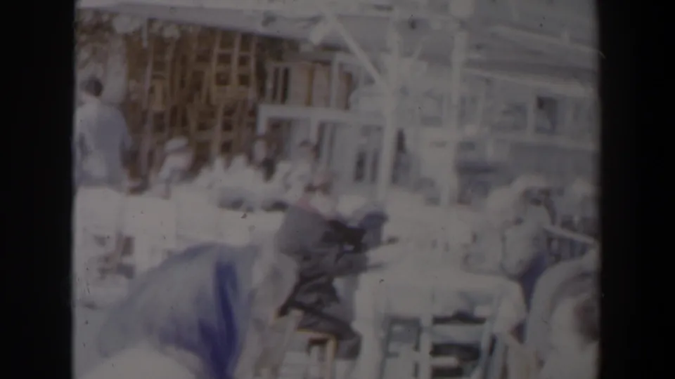 Faded 8mm frame of a communal seating area in Israel, showing adults at tables with chairs and a bright structural backdrop.