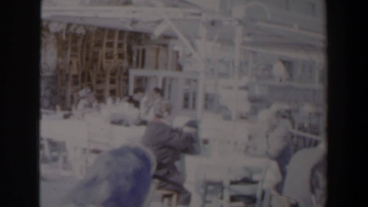 Overexposed outdoor restaurant scene with seated patrons and harbor structures faintly visible.
