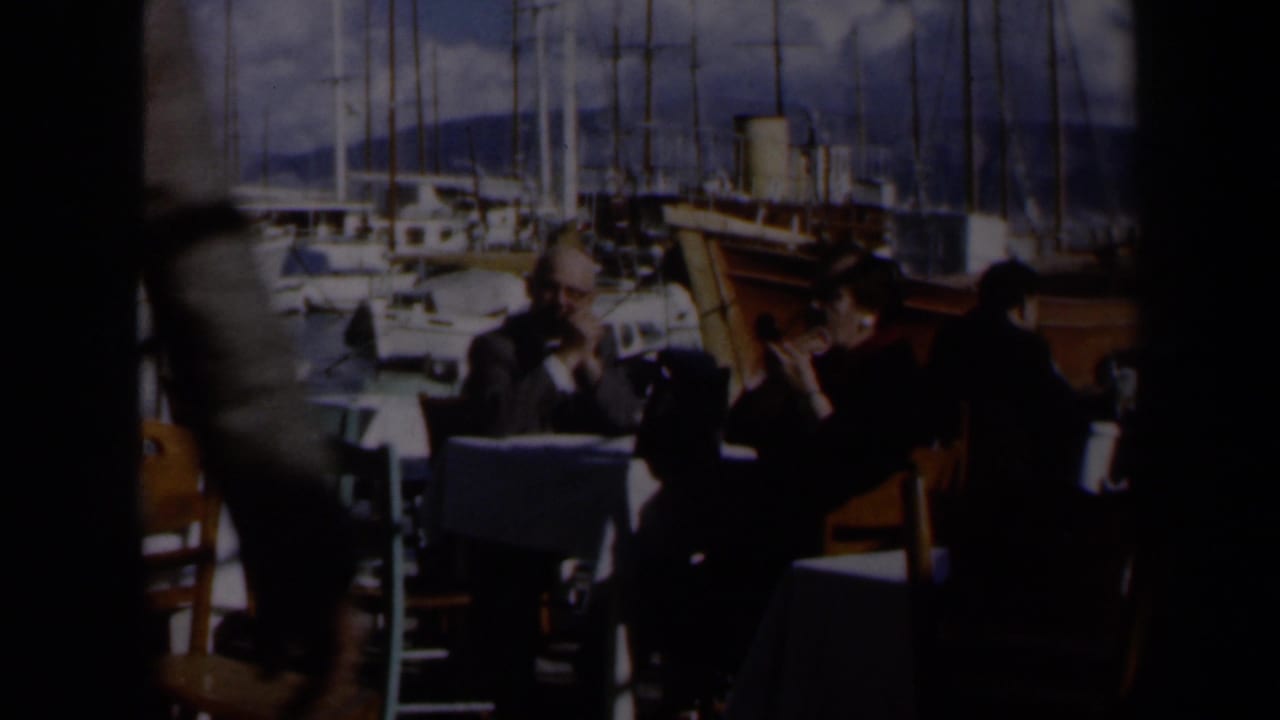 A man and woman sit at a seaside restaurant table in Israel, with moored boats, tall masts, and hills in the background.
