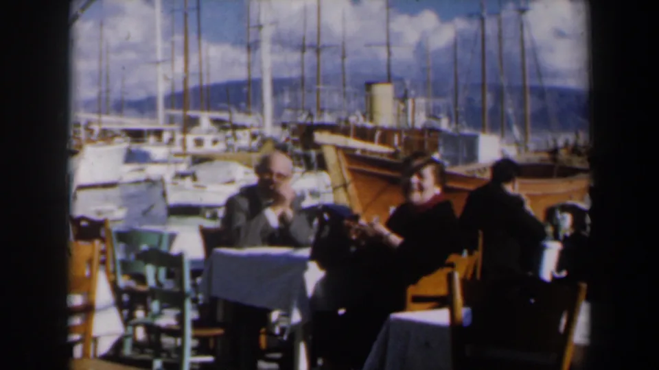 In 1960 Israel, a seated man and woman converse at an outdoor restaurant table beside a marina crowded with masts.