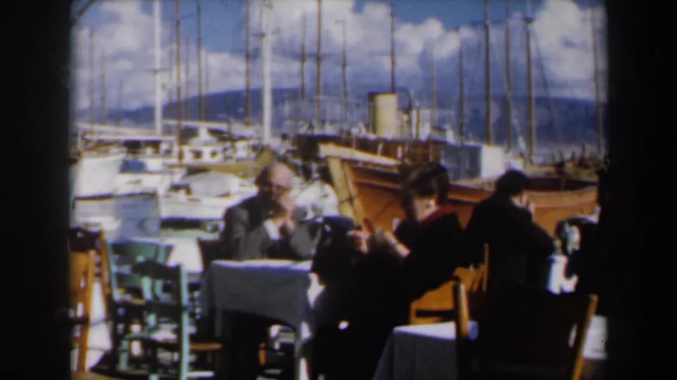 A 1960 Israel harbor restaurant frame shows adults seated at tables, with boats, masts, blue sky, and hills beyond.