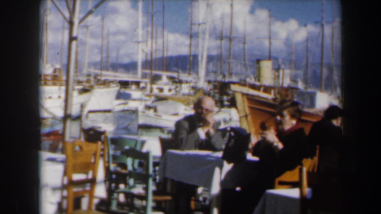 Adults seated at an outdoor restaurant table beside a marina with sailboats behind them.