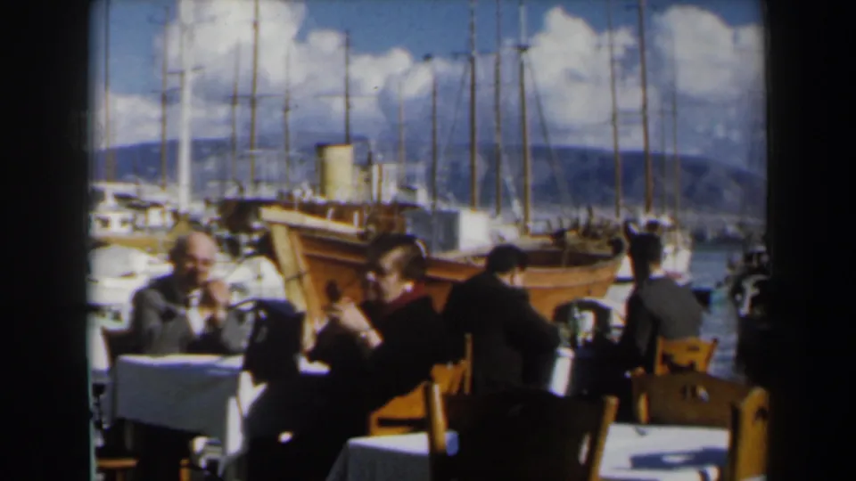 1960 Israel frame of people eating and socializing outdoors near a marina, with boats and hills across the water.