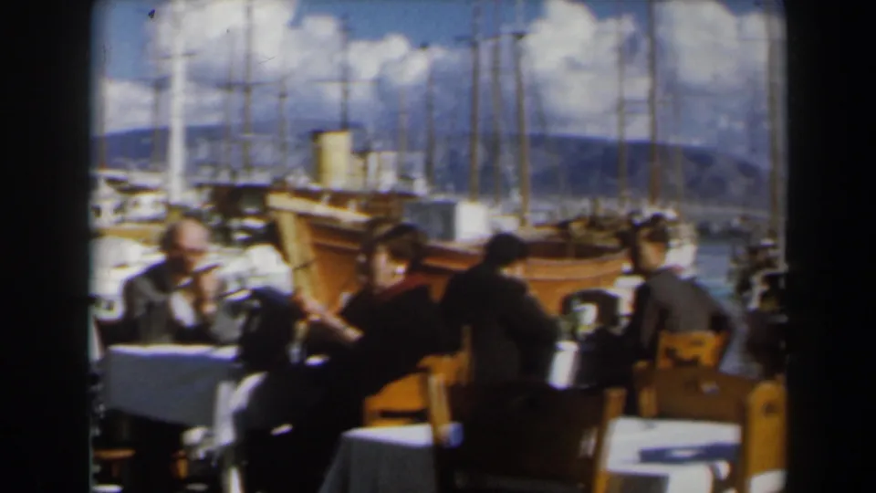Adults at waterfront restaurant tables in Israel, framed by a wooden boat, sailboat masts, bright sky, and distant hills.