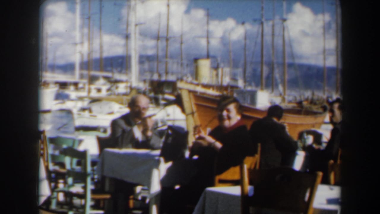 1960s Israel home-movie frame of adults dining beside a marina with sailboats, blue sky, and distant hills in the background.