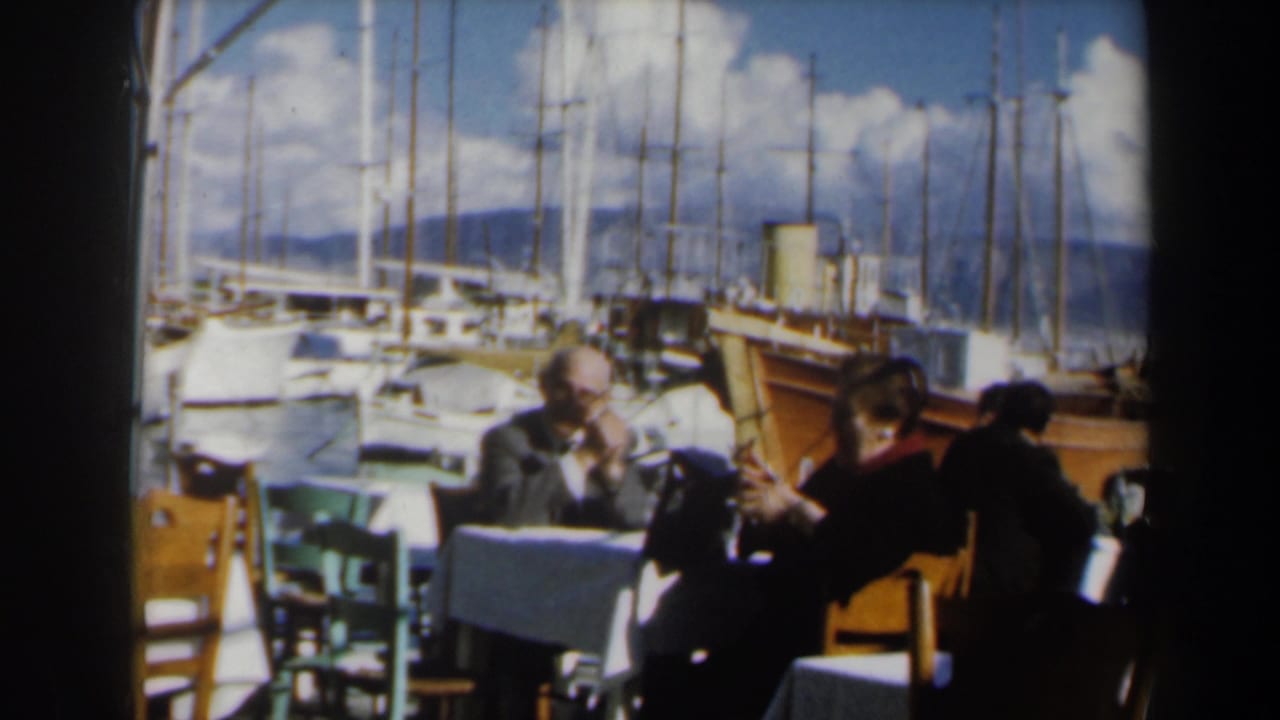 Outdoor diners sit near a wooden boat with sailboat masts filling the background.