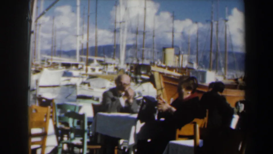 Adults at a waterfront restaurant in Israel, 1960, with a wooden boat, marina masts, clouds, and hills behind the table.