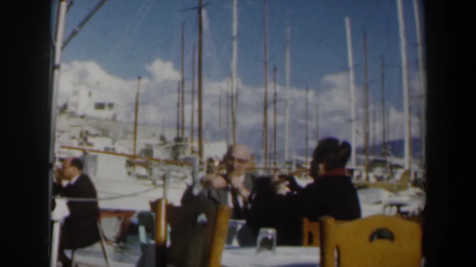 1960s Israel marina dining scene with seated adults, cafe chairs, moored boats, tall masts, bright clouds, and distant hills.